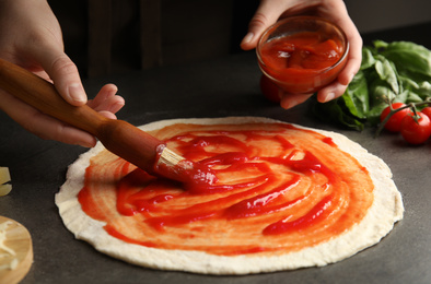 Woman spreading tomato sauce onto pizza crust at grey table, closeup