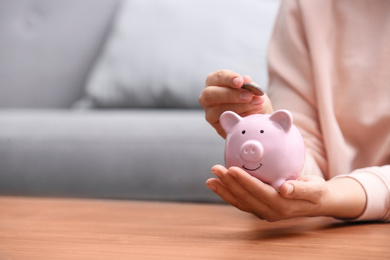 Woman putting coin into piggy bank at wooden table, closeup. Space for text