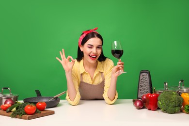 Young housewife with glass of wine, vegetables and different utensils on green background