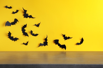 Empty grey table near yellow wall decorated with paper bats, space for text. Halloween celebration