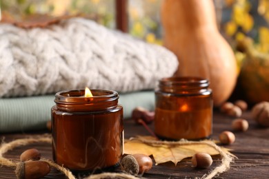 Burning scented candles, warm sweaters and acorns on wooden table near window, closeup. Autumn coziness