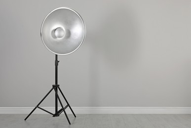 Professional beauty dish reflector on tripod near grey wall in room, space for text. Photography equipment