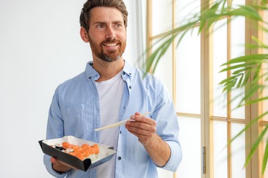 Happy man eating sushi rolls with chopsticks indoors