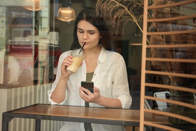 Young beautiful woman with smartphone drinking coffee in cafe, view through window glass