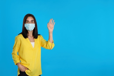 Photo of Young woman in protective mask showing hello gesture on light blue background, space for text. Keeping social distance during coronavirus pandemic