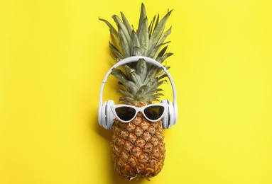 Photo of Pineapple with sunglasses and headphones on yellow background, top view. Creative concept