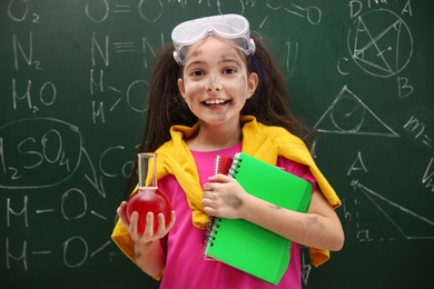 Schoolgirl holding flask and notebooks near chalkboard with chemical formulas