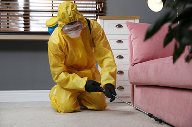 Pest control worker in protective suit spraying insecticide under sofa at home