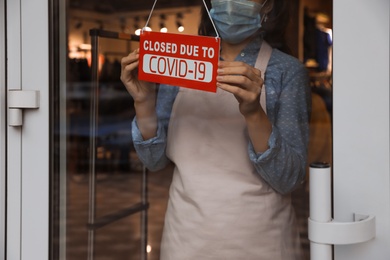 Woman in mask putting red sign with words "Closed Due To Covid-19" onto glass door, closeup