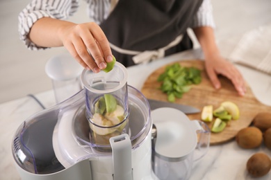 Young woman putting fresh kiwi into juicer at table in kitchen, closeup