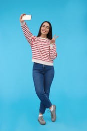 Photo of Smiling young woman taking selfie with smartphone and showing peace sign on light blue background