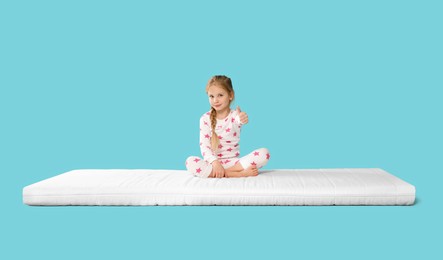 Photo of Little girl sitting on mattress and showing thumb up against light blue background