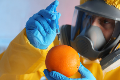 Scientist in chemical protective suit injecting orange against color background, focus on fruit