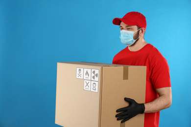 Courier in mask holding cardboard box with different packaging symbols on blue background. Parcel delivery