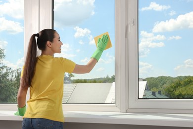 Happy young woman cleaning window glass with sponge cloth indoors