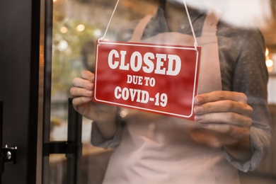 Woman putting red sign with words "Closed Due To Covid-19" onto glass door, closeup