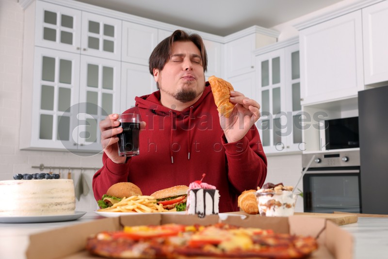 Overweight man eating tasty croissant at table in kitchen