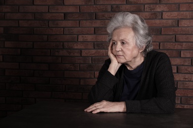 Poor upset woman sitting at table near brick wall. Space for text