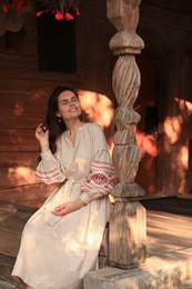 Photo of Beautiful woman in embroidered dress sitting near carved wooden column on terrace. Ukrainian national clothes