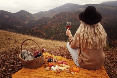 Young woman with glass of wine having picnic in mountains on autumn day, back view