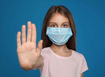 Little girl in protective mask showing stop gesture on blue background. Prevent spreading of coronavirus