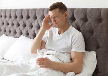 Young man with terrible headache holding pills and glass of water in bed