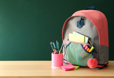 Stylish backpack and different school stationery on wooden table near green chalkboard, space for text. Back to school