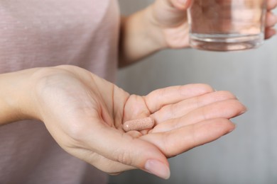Woman holding gelatin capsule and glass of water on grey background, closeup