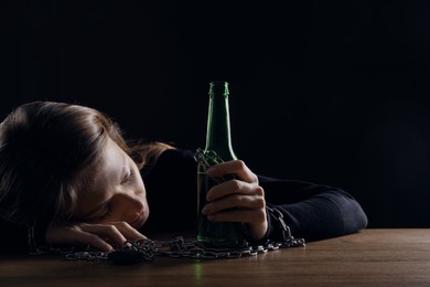 Alcohol addiction. Woman chained with bottle of beer sleeping at wooden table against black background