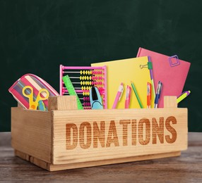 Donation box with different school stationery on wooden table near chalkboard