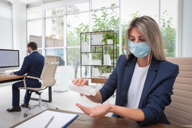 Photo of Office employee in mask applying hand sanitizer at workplace