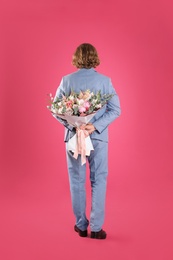 Man in stylish suit hiding beautiful flower bouquet behind his back on pink background