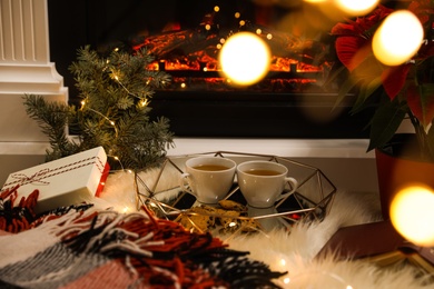 Photo of Cups of hot drink and cookies near fireplace indoors