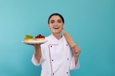 Photo of Happy professional confectioner in uniform with delicious tartlets and rolling pin on light blue background