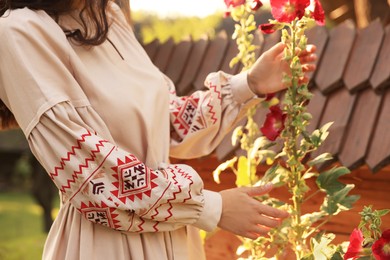 Woman wearing beautiful embroidered dress in blooming garden on sunny day, closeup. Ukrainian national clothes