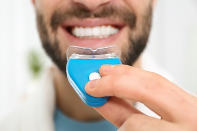 Young man using teeth whitening device on light background, closeup