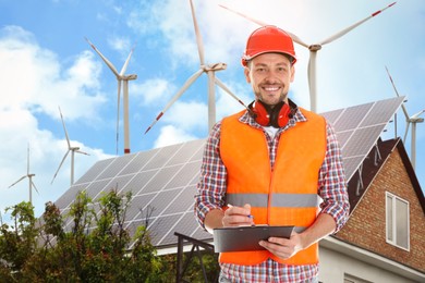 Image of Industrial engineer in uniform and view of wind energy turbines near house with installed solar panels on roof