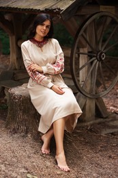 Beautiful woman wearing embroidered dress sitting near old wooden well in countryside. Ukrainian national clothes