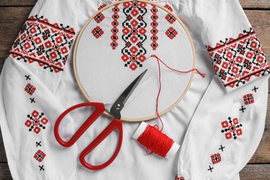 Photo of Shirt with red embroidery design in hoop, needle, scissors and thread on wooden table, top view. National Ukrainian clothes