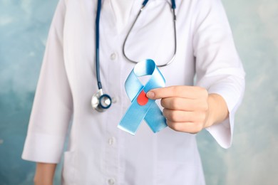 Photo of Doctor holding light blue ribbon with paper blood drop on color background, closeup. World Diabetes Day
