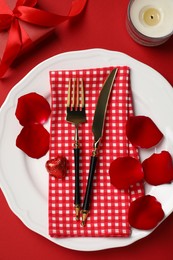 Photo of Beautiful table setting with gift box and rose petals on red table for romantic dinner, flat lay