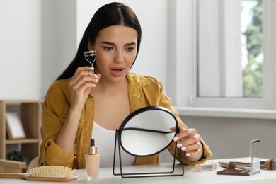 Emotional young woman using eyelash curler while doing makeup at table indoors