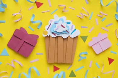 Photo of Birthday party. Paper cupcake, gifts and confetti on yellow background, flat lay