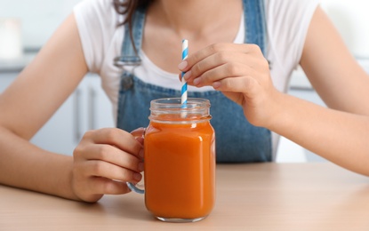 Woman with mason jar of tasty carrot juice at table indoors, closeup
