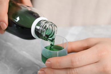 Photo of Woman pouring syrup from bottle into measuring cup at grey marble table, closeup. Cold medicine