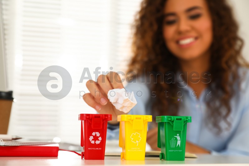 Young African-American woman throwing paper into mini recycling bin at table in office, focus on hand