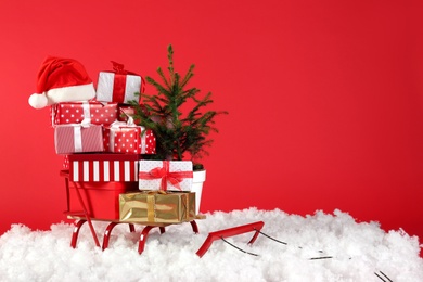 Sleigh with presents, Santa hat and fir tree in artificial snow on red background. Space for text