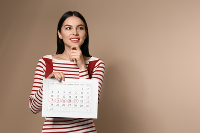 Pensive young woman holding calendar with marked menstrual cycle days on beige background. Space for text