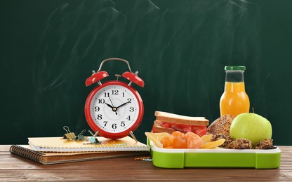 Lunch box with appetizing food and alarm clock on wooden table near green chalkboard