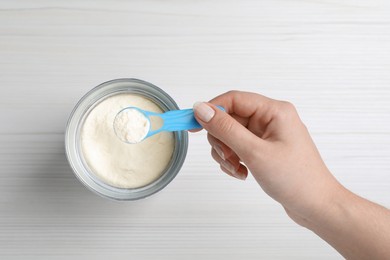 Woman taking powdered infant formula with scoop from can at white wooden table, top view